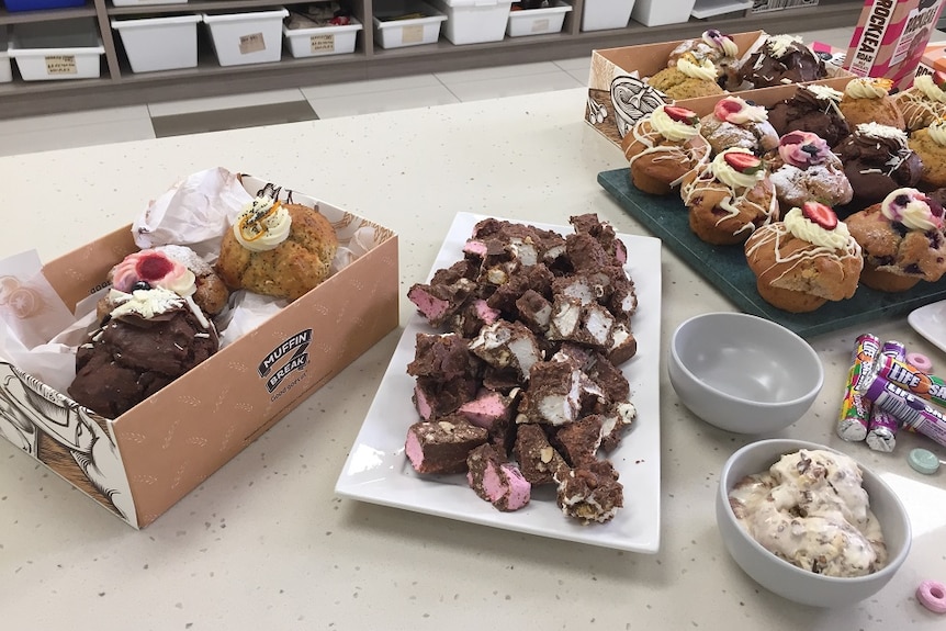 Different muffins in Muffin Break boxes, next to a platter of rocky road, a bowl of ice cream and life saver lollies