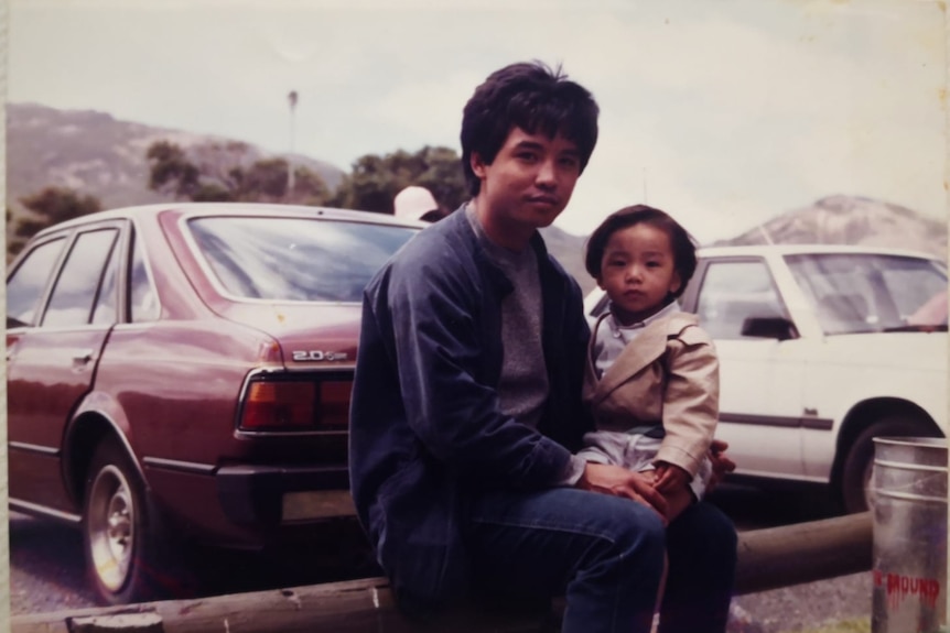 An old photo of a father and son posing near 1970s cars.