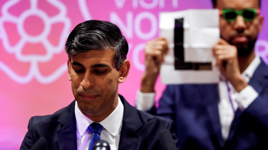 Rishi Sunak looks down as he speaks while a man stands behind him holding a sign that says: "L".