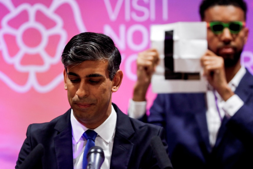 Rishi Sunak looks down as he speaks while a man stands behind him holding a sign that says: "L".