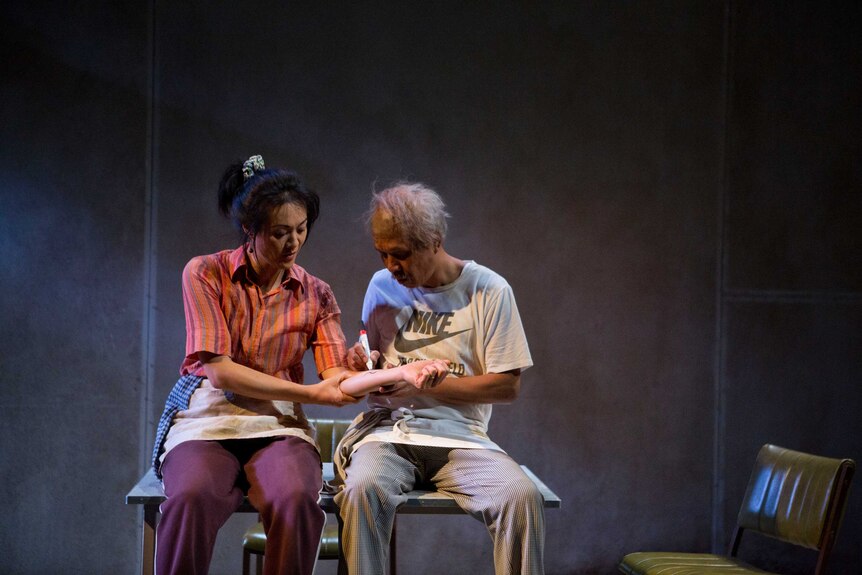 A woman and older man sit on a table, the man is drawing on the woman's arm in texta. Both wear aprons.