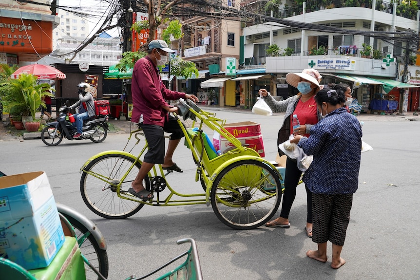 A man on a bike talking to two women holding plastic bags of food 