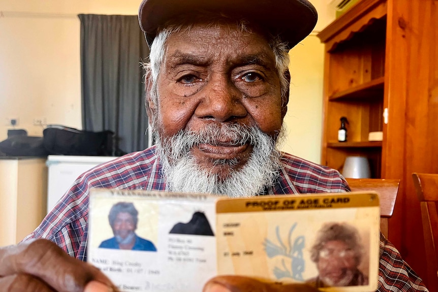 An Aboriginal man with a white beard holds up his ID cards.