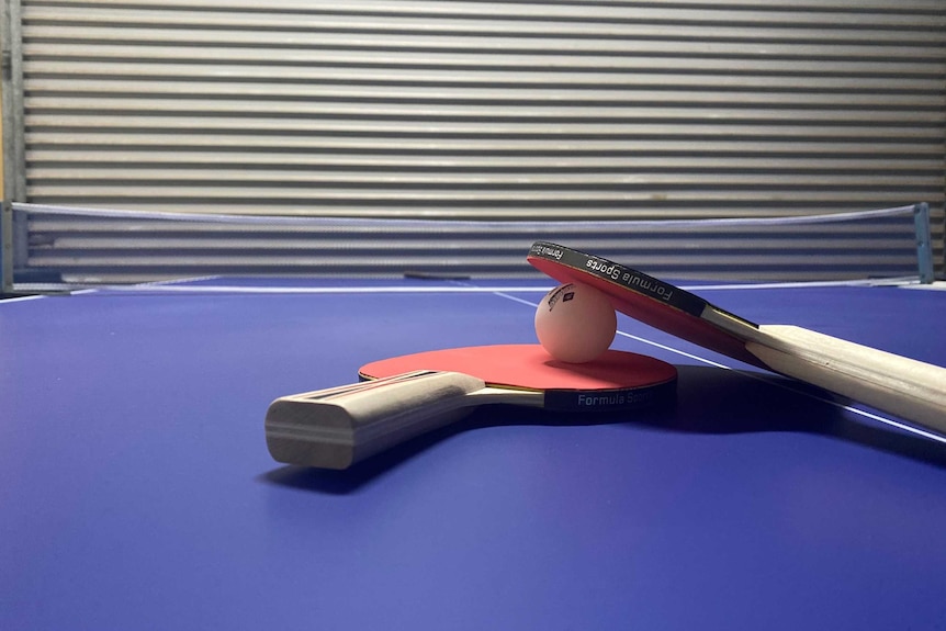 Two table tennis racquets and a ball lying on a blue table inside a room.