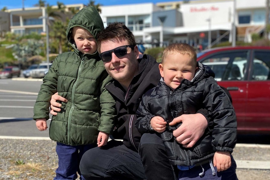 A man squatting while hugging two young boys near a carpark