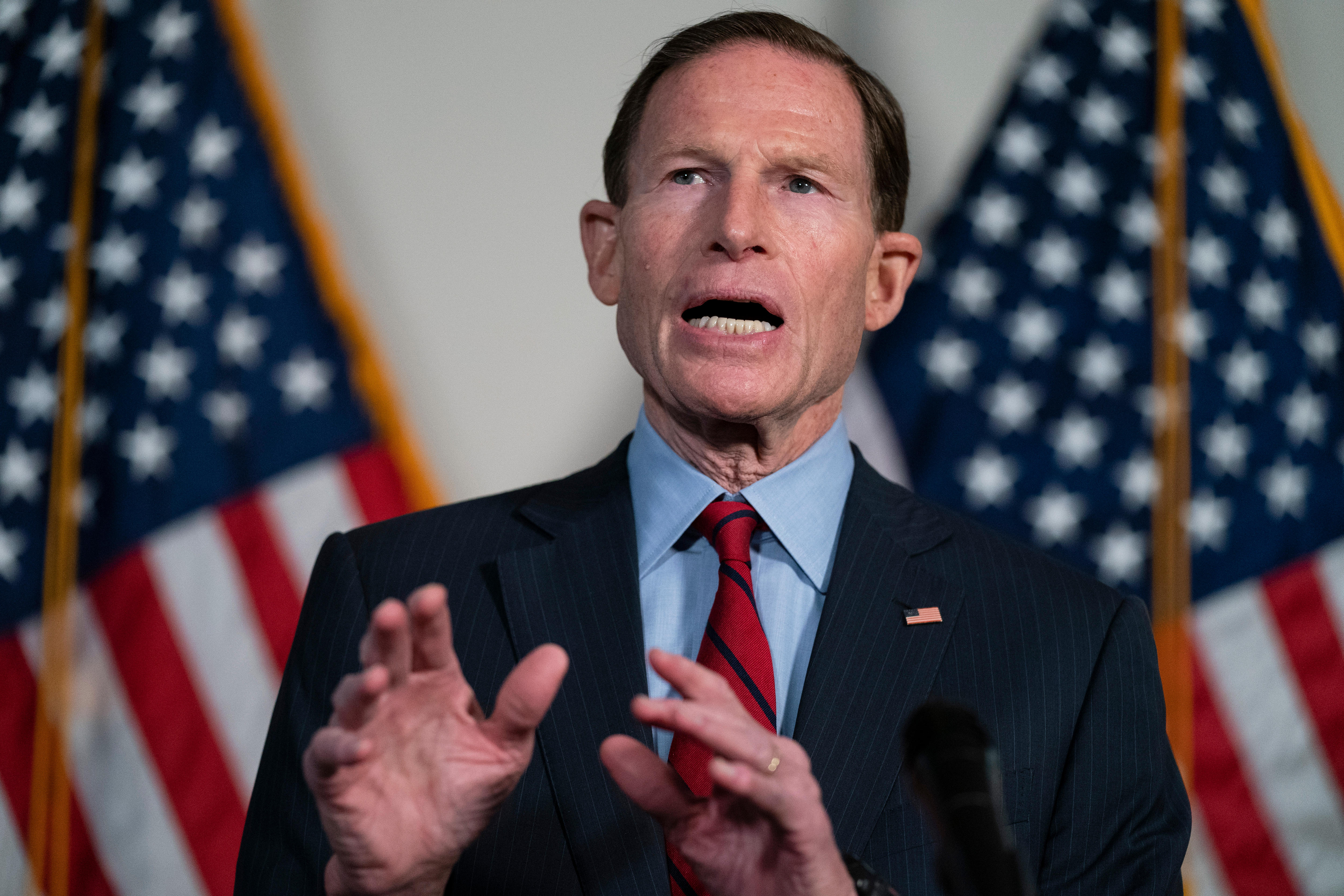 a man in a navy blue suit and red tie holds his hands in front of him while giving a speech