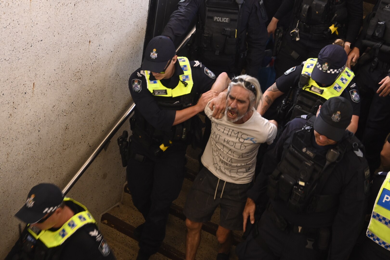 A man in a white shirt with writing on it being escorted by police  