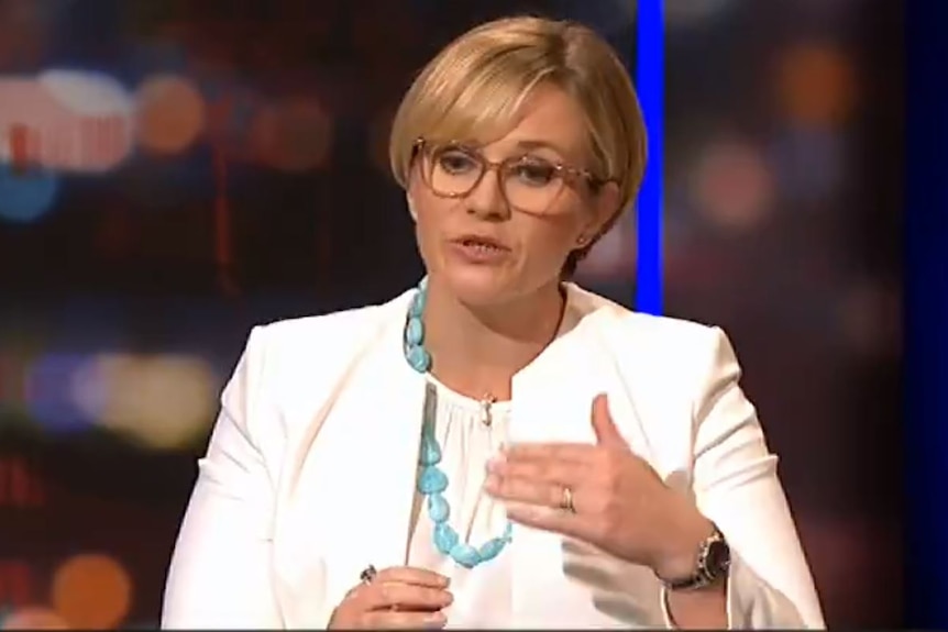 A woman with short blond hair and glasses wears a white jacket and blouse.