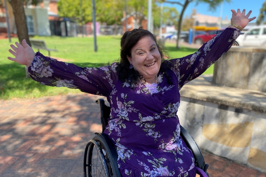 A woman in a purple dress smiles widely with her arms stretched out