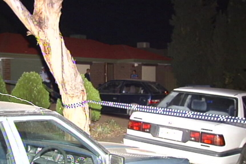 Blue and white police tape borders a tree and white car, with a red brick house with people outside