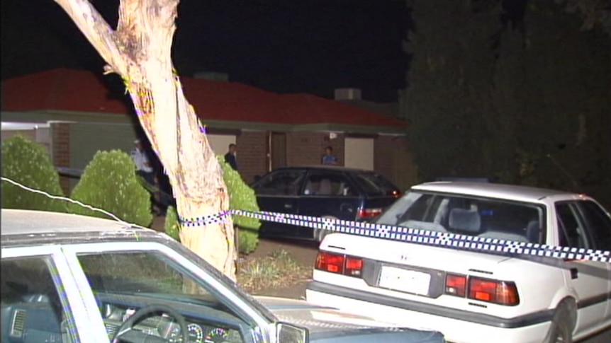 Blue and white police tape borders a tree and white car, with a red brick house with people outside