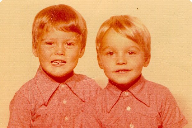 A faded colour photo of two young boys sitting together. They have matching shirts, but one has darker hair than the other