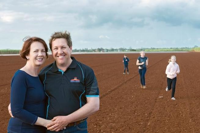 A couple stands in the foreground of a red dirt paddock with three children running toward the camera in the background