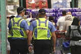 Two police officers with yellow high-viz vests walk through a supermarket surrounded by shoppers.