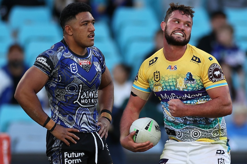 A Gold Coast Titans NRL player holds the ball with his right hand after scoring a try as a Canterbury opponent looks on.