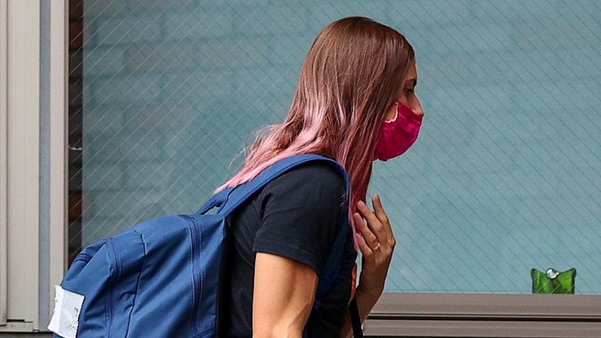 A woman with dyed pink hair wearing a backpack and face mask walks into a building.