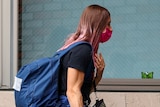 A woman with dyed pink hair wearing a backpack and face mask walks into a building.