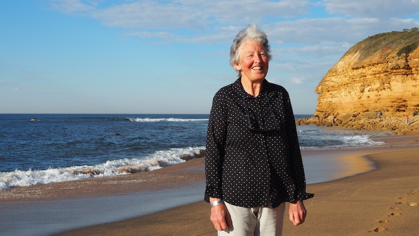 A woman in a black long sleeve shirt with white spots stands on a beach with a cliff in the background and laughs.