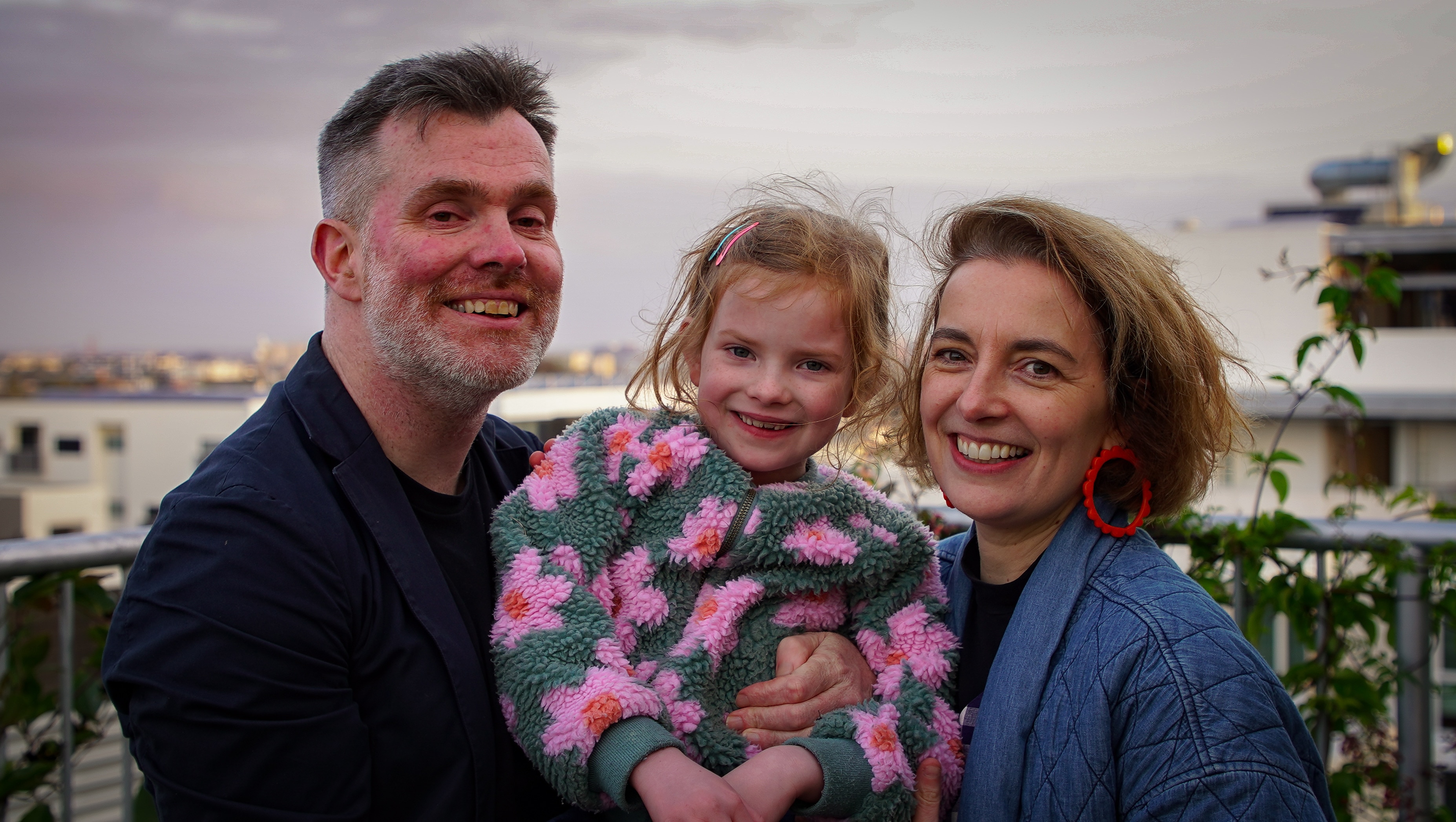 A mother and father holding their young daughter between them, smiling. They are standing on an apartment balcony.