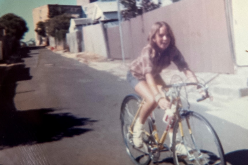 1970s image of a girl in denim shorts and a checked shirt riding a road bike down a suburban alleyway