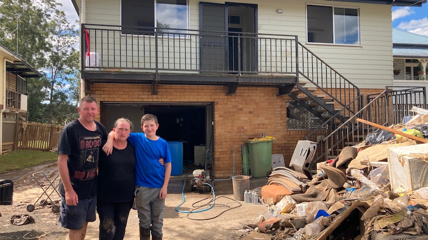 Man in black shirt, woman in black shirt, teenage boy in blue shirt standing next to pile of rubbish in front of 2 storey house