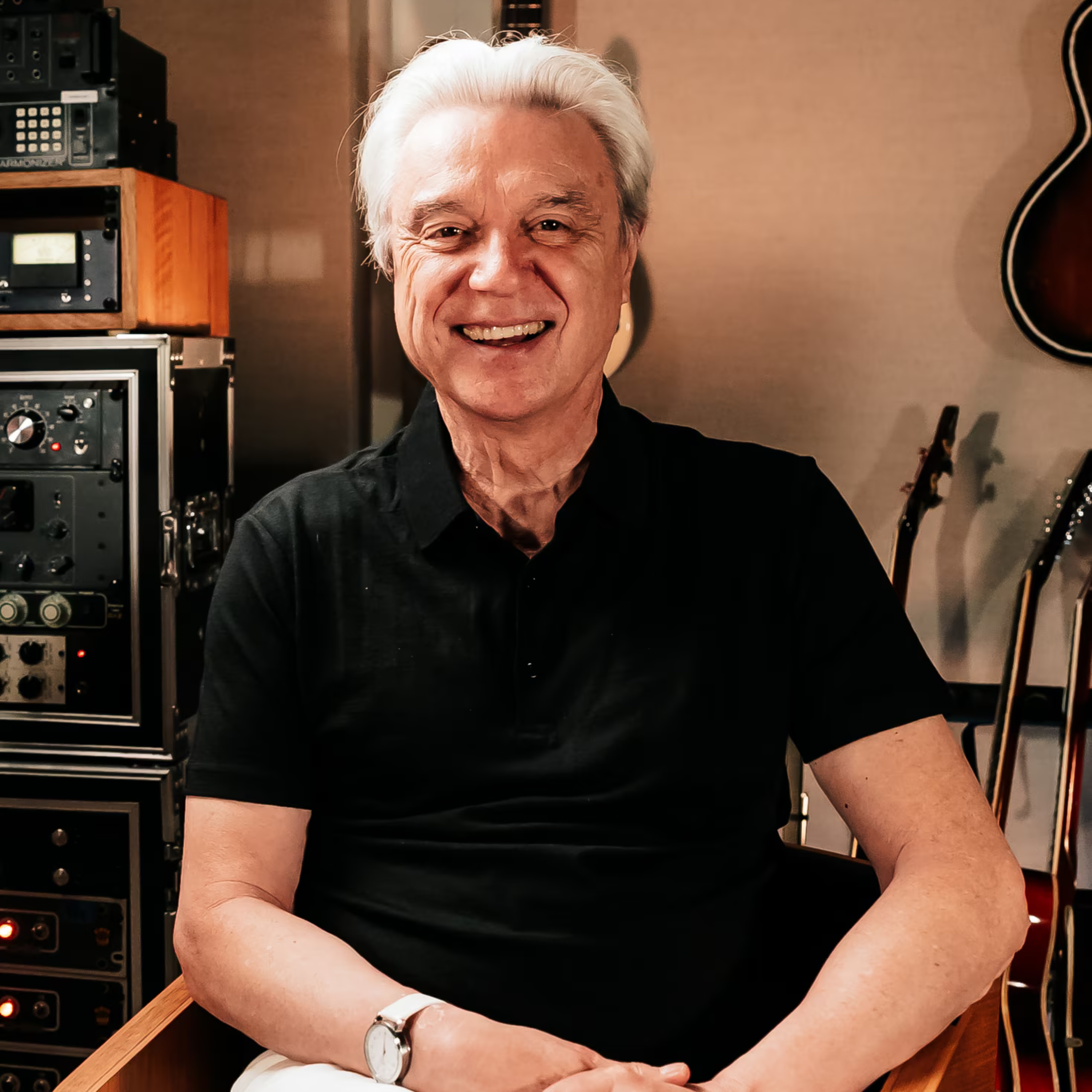 Smiling man with white hair in a black shirt sits in a recording studio, surrounded by audio equipment and guitars