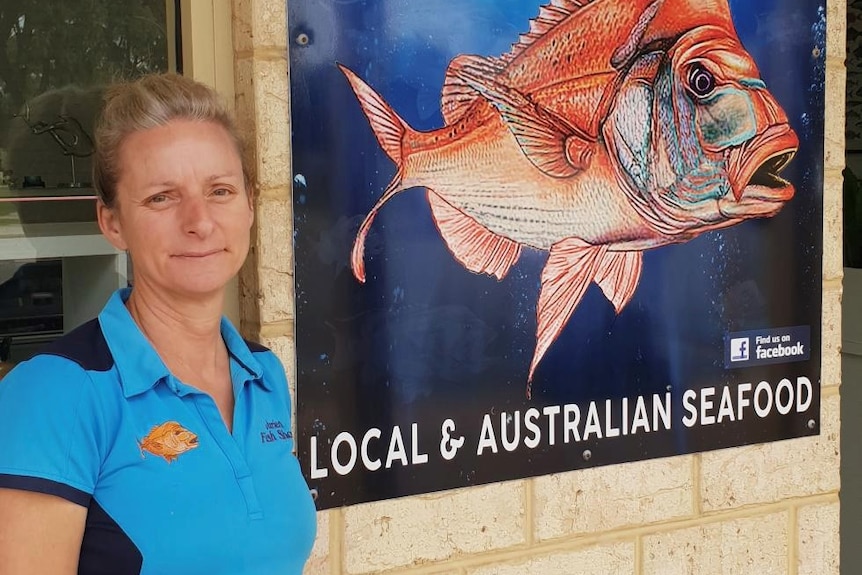 A woman stands looking sadly at the camera near a sign for her fish shop