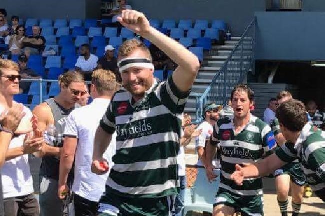 Rugby player smiles at camera as he runs onto field with arm raised.