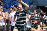 Rugby player smiles at camera as he runs onto field with arm raised.