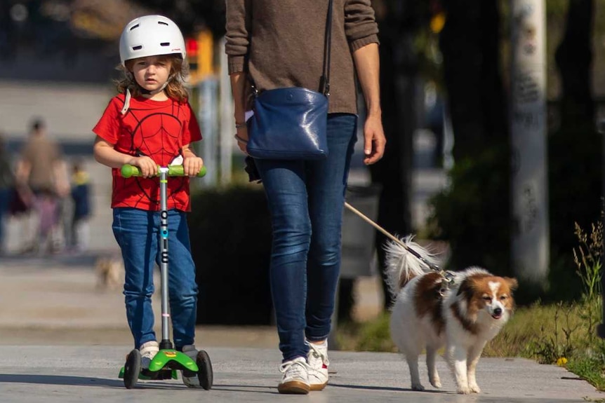 A group of children and adults, and one dog, ride scooters and skateboards down a pathway in sunny Barcelona.