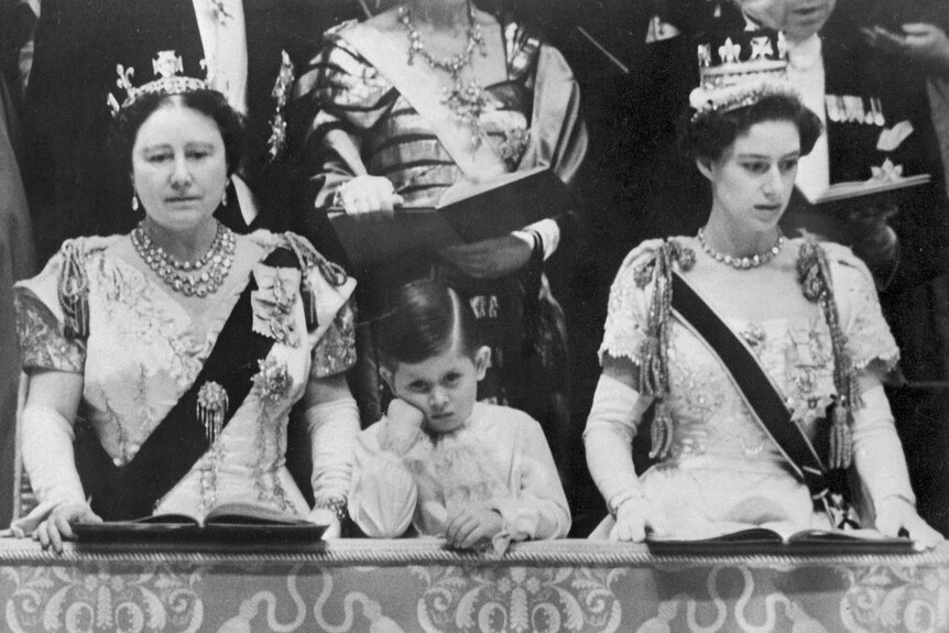 King Charles as a child leans elbow on a chair and head on his hand, looking bored, next to Queen Mother and Princess Margaret
