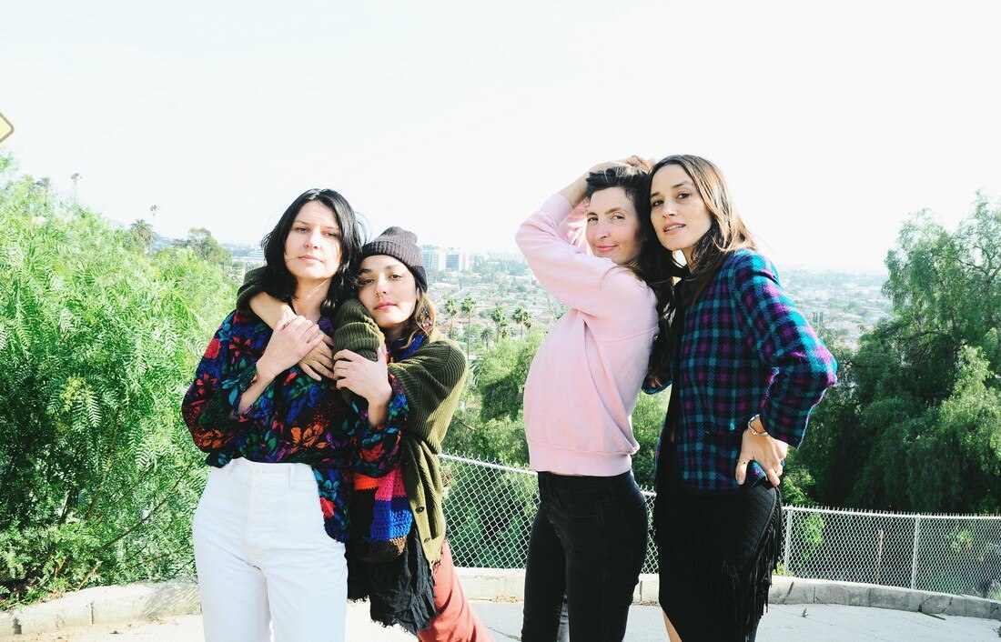 Two sets of two women, members of the band Warpaint, stand arm in arm in an outdoor area