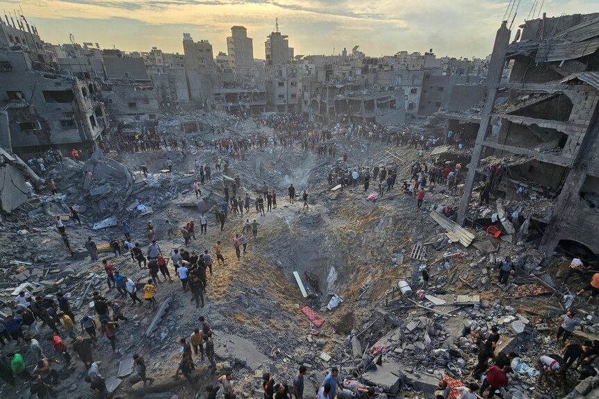 People crowd around a huge crater in a decimated urban area. 