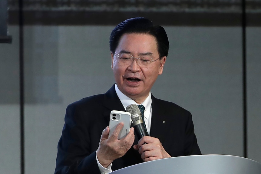 A middle-aged Taiwanese man in a suit looks at his phone while standing behind a podium.