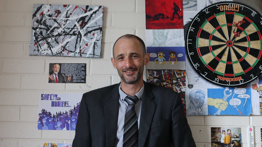 Man in a suit smiling in front of a cream-coloured wall, covered in posters and a dart board.