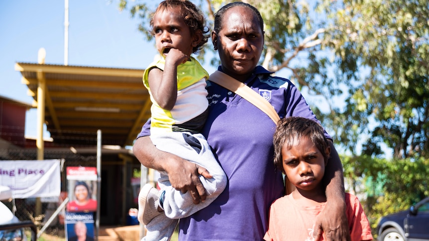 A woman stands in the remote community of Binjari with two children. Election corflutes are in the backdrop