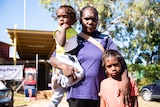 A woman stands in the remote community of Binjari with two children. Election corflutes are in the backdrop