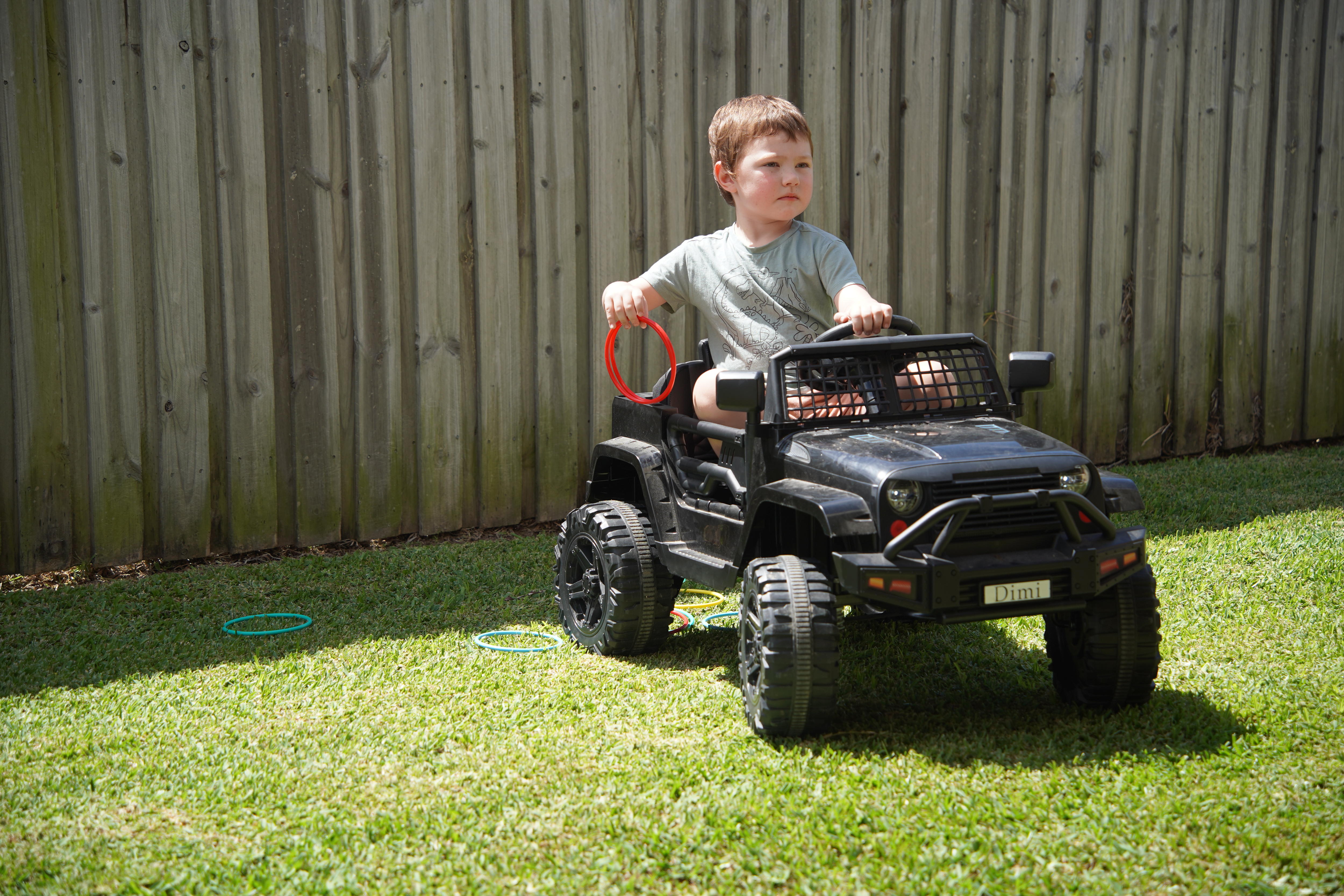 A young boy sits on a toy car in a backyard with one hand on the steering wheel.