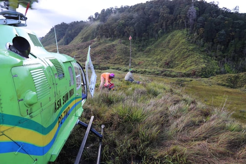 A helicopter sits on top of a mountain as two people are seen in the distance.