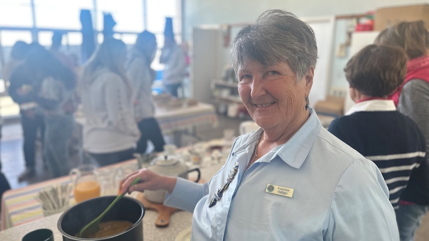 A grey-haired woman smiles as she serves soup from a large pot. 
