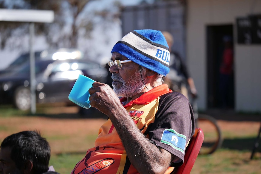 An Indigenous man wearing rugby league clothes sips from a plastic cup as he watches a match out of frame.