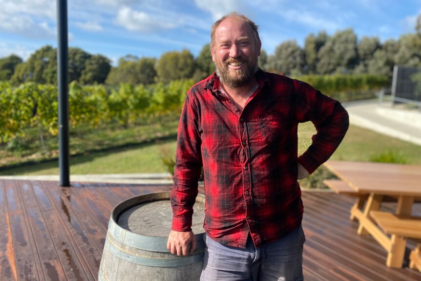 A man in a checkered shirt leans on a wine barrel, smiling