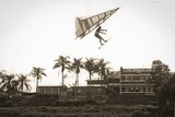 Man flying a hang glider past on a river with an old brick hotel building in the background.