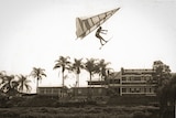 Man flying a hang glider past on a river with an old brick hotel building in the background.