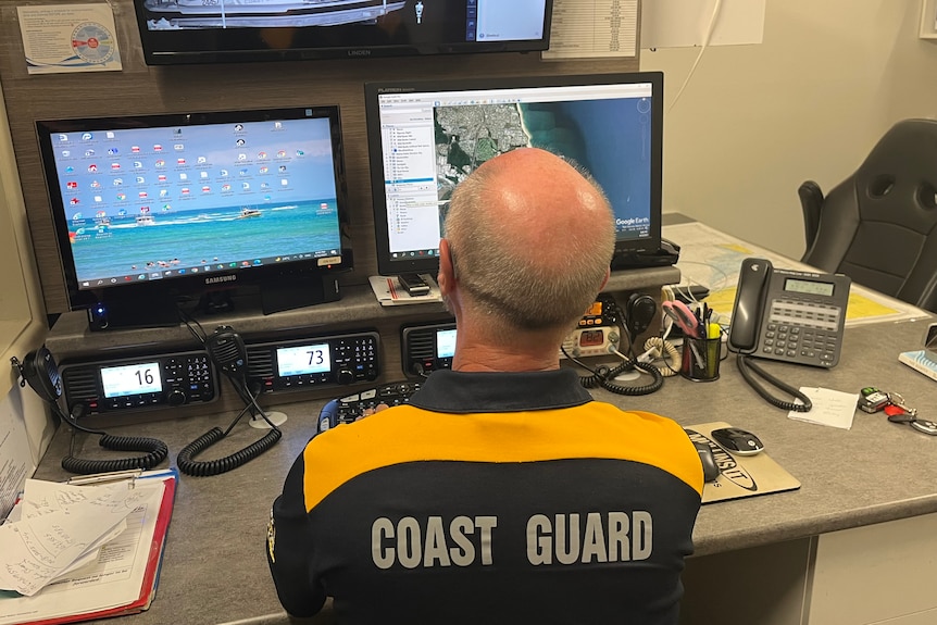 A middle-aged bald man wearing a golf shirt that says 'coast guard' sits in office and looks at monitors