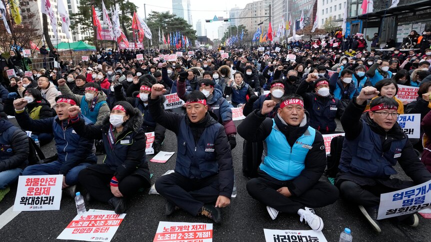 Thousands of people sit on a main road in front of signs demanding better working conditions during a protest.