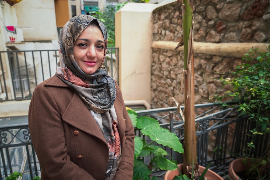 A woman wearing a patterned hijab and brown jacket smiles in a garden.