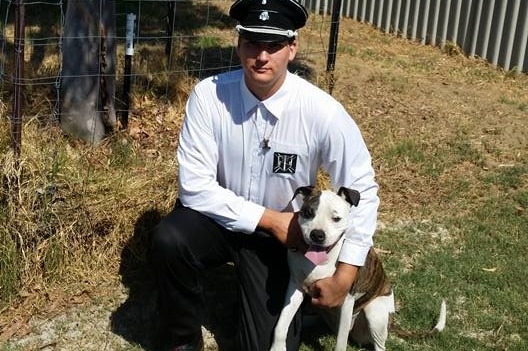 A man wearing a white and black Neo_nazi outfit kneels next to a dog in the yard of a house.