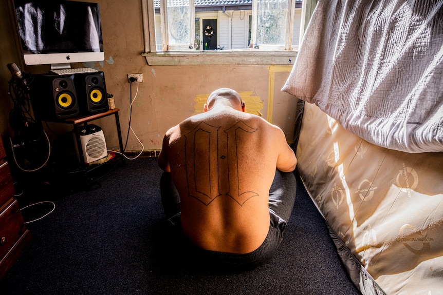 A shirtless man with a large tattoo of "OC" on his back sits on a bedroom floor, his mattress against the wall.