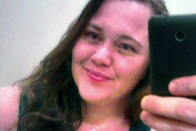 Cara Lee Hall, with long brown hair, wearing a black singlet and a small cross around her neck, smiles taking a mirror selfie.